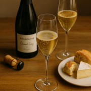 Table en bois clair avec deux flûtes de champagne d’auteur pétillant, une bouteille élégante en arrière-plan et une assiette de fromage et pain rustique